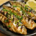 Grilled mackerel fish with chopped green onions, sesame seeds, and lemon wedges on a black plate, showcasing a savory Asian-inspired seafood dish.